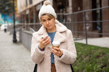Genç, güzel bir kadının cep telefonu kullanan portresi, mesaj atan, geri dönüşümlü bardağı tutan, sıcak kahve içen, çay içen. Şapka, kürk manto giyiyor. Sokak tarzı.