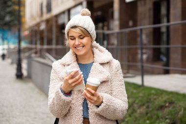 Genç güzel bir kadının cep telefonu kullanan portresi, mesaj atan, geri dönüşümlü bardağı tutan, sıcak kahve içen, çay içen. Gülümsüyor, şapka takıyor, kürk manto giyiyor. Sokak tarzı. Keyfiniz yerinde, mutlu insanlar..