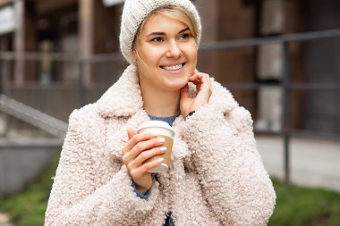 Genç ve güzel bir kadının portresi geri dönüşümlü bardağı elinde sıcak kahve ya da çayla elinde tutuyordu. Gülümsüyor, şapka takıyor, kürk manto giyiyor. Sokak tarzı. Harika bir ruh hali, mutlu insanlar. Sokak tarzı, şehir hayatı.