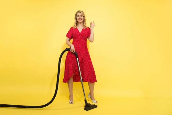 Positive blonde woman on yellow background, red dress, with vacuum cleaner in hands smiles, points with hand, empty space for advertising, studio shot, concept of cleaning service for homes, offices.