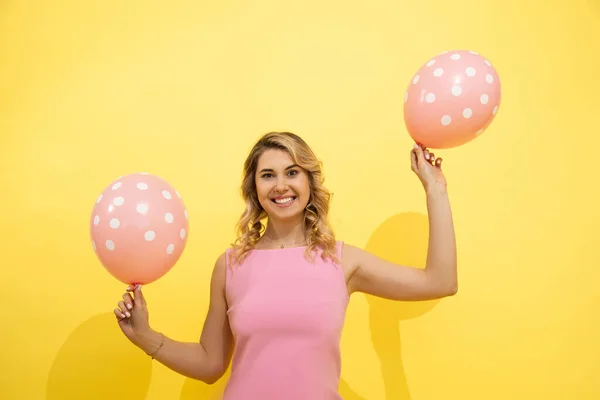 ピンクの風船を手にしたポジティブな若い長髪のブロンドの女性が笑顔で 遊び心のある雰囲気の中でカメラを見ています 休日の概念は 黄色の背景に撮影スタジオ — ストック写真