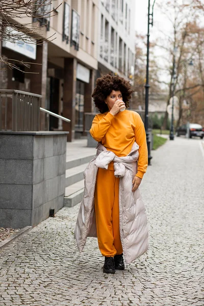 Genç, güzel beyaz kadın portresi, 40 yaşında spor kıyafetleri, sokakta yürüyen, tek kullanımlık kahve bardağı. Afro saç stili. Sonbahar gününün tadını çıkarıyorum. Neşeli insanlar, şehir hayatı