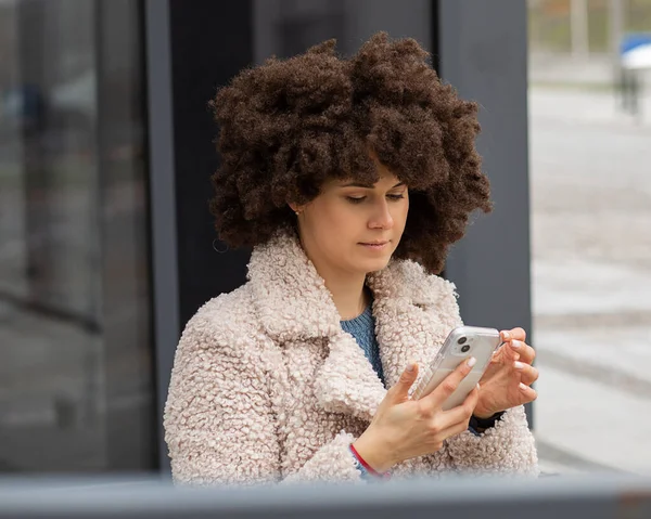 Urban style. Young modern white-skinned, beautiful woman. With hair Type Afro hair Style. Lush hairstyle uses mobile phone for text message. Portrait in city against background of concrete building.