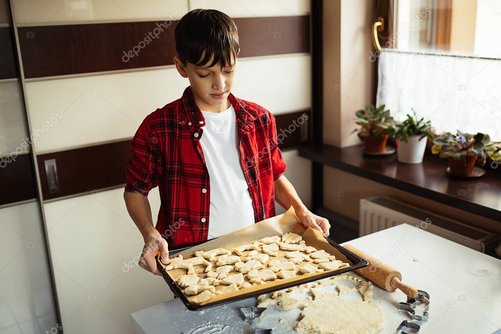 Boy teenager in checkered shirt in red, holds in his hands Baking sheet covered with parchment with cookies. Christmas cut-off biscuits. Animal shape. Heading to oven.