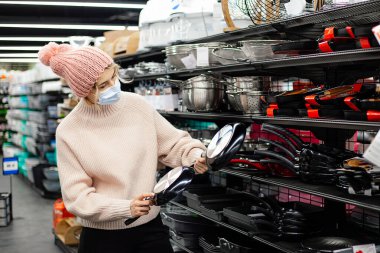 Koruyucu maskeli ve örgü şapkalı bir kadın süpermarketteki mutfak gereçleri bölümünde tavayı dikkatlice seçiyor. Salgın hastalık alışverişi. Yemek için tabaklar. Ev hayatı. Her gün endişeleniyorum. Yaşam biçimi.