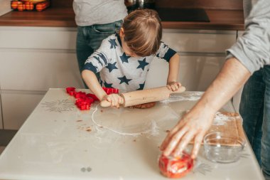 Ev pijamalarına tutkulu sevimli çocuk kurabiye pişirmek için masaya raptiye takmış hamuru yuvarlıyor. Babam ve annem onun yanında duruyor. Mutlu aile, takım çalışması, çocuklar için pişirme dersleri..