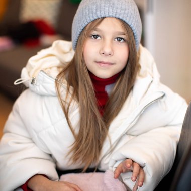 Gri örgü şapkalı ve uzun saçlı kameraya bakan beyaz ceketli kızın portresi. Mutlu bir çocukluk. Tatiller ve seyahatler. Güzel çocuklar. Olumlu duygular. Yaşam Tarzları.