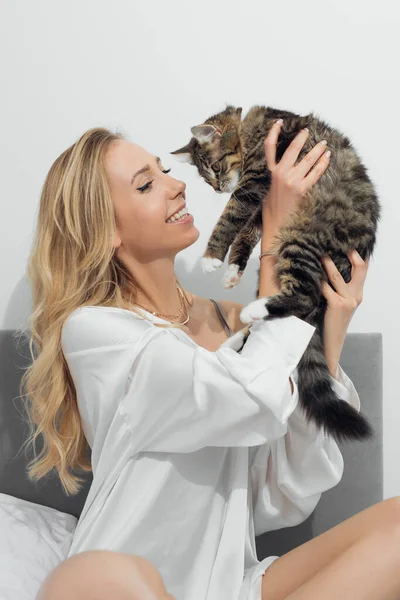 Egy fiatal szőke nő portréja, barna fehér és fekete csíkos macskával a kezében. Egymásra nézünk. Gyengéden ölelő cica — Stock Fotó