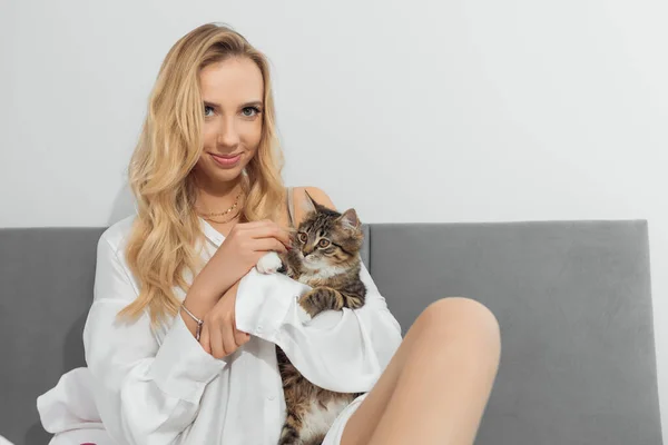 Retrato de una joven rubia sosteniendo un gato marrón blanco y negro mirando a la mano. Alimentar al gato. Gatito abrazando suavemente —  Fotos de Stock