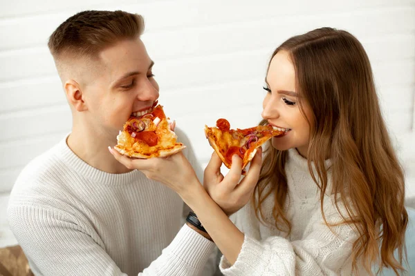 Desfrute de pizza.Jovem mulher e seu amante estão felizes, almoçando juntos e brincando alimentando uns aos outros, eles estão sentados contra a parede branca. Encontro em casa. Relações românticas. — Fotografia de Stock