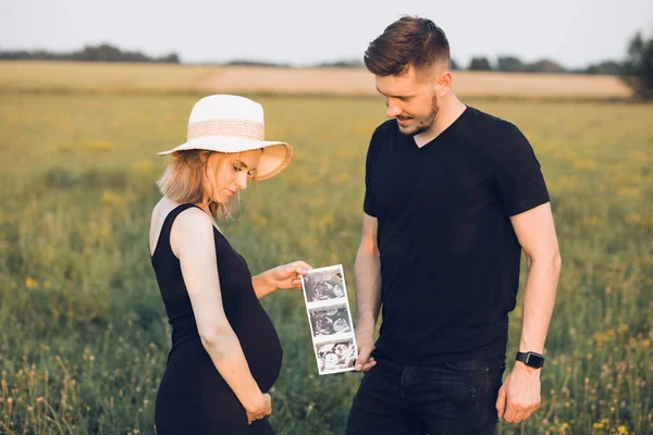Νεαρή έγκυος γυναίκα και σύζυγος περπατούν στο χωράφι, κρατούν φωτογραφίες από υπέρηχο. Ζευγάρι περιμένει μωρό, χαλαρώστε έξω — Φωτογραφία Αρχείου
