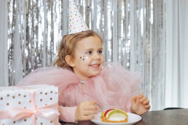 Kısa kıvırcık saçlı, yüzünde pembe kabarık elbisesi olan ve meyveli pasta ve hediye kutusunun önünde şapkalı gülümsemesi olan küçük bir kız.