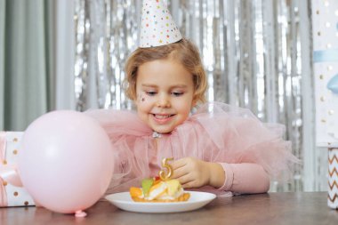 Kıvırcık saçlı, yüzünde pembe kabarık elbiseli ve şapkalı beş numaralı mumla meyveli pastaya bakan küçük bir kız..