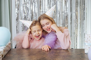 İki güzel kıvırcık saçlı, yüzünde kabarık elbiseli ve doğum günü şapkalı dillerini gösteren iki güzel kız..