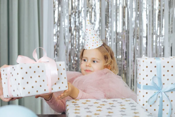Little girl with short curly hair and stars on face in pink poofy dress and cap taking gift box wrapped in white paper. — Stock Photo, Image