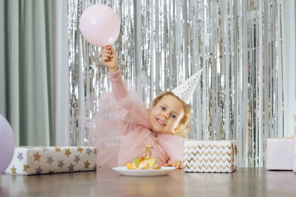 Roztomilé malé usmívající se dívka s krátkými kudrnaté vlasy a hvězdy na tváři v růžové poofy šaty výtah růžový balón v blízkosti ovoce dort. — Stock fotografie