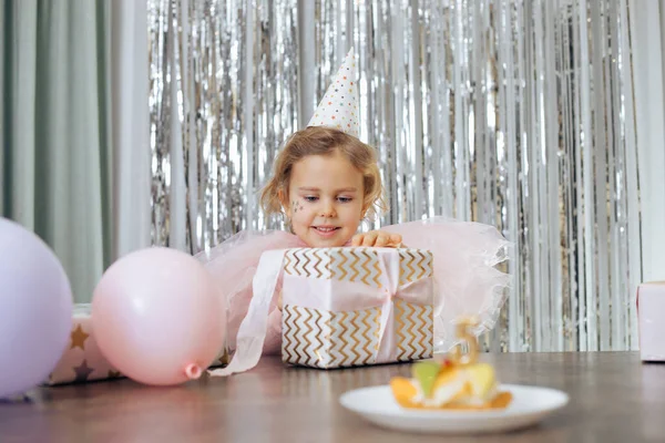 Carino poco sorridente ragazza con i capelli corti ricci e le stelle sul viso in abito rosa poofy, cap scartare confezione regalo vicino palloncini. — Foto Stock