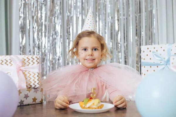 Cute little girl z krótkimi kręconymi włosami i gwiazdami na twarzy w różowy peofy sukienka i czapka wygląd uśmiechnięty przed ciastem owocowym — Zdjęcie stockowe