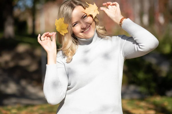 Bella donna sorridente di mezza età con lunghi capelli biondi ondulati in maglione roll-neck tenere foglie d'acero gialle vicino alla testa. — Foto Stock
