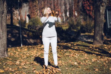 Uzun dalgalı sarı saçlı, yuvarlak yakalı kazaklı, gülümseyen orta yaşlı kadın kusmak için sarı akçaağaç yaprağı yığını tutuyor..