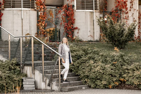 Atractiva mujer de mediana edad con chaqueta a cuadros y suéter gris con cuello enrollable que baja escaleras de hormigón en el día de otoño.. —  Fotos de Stock