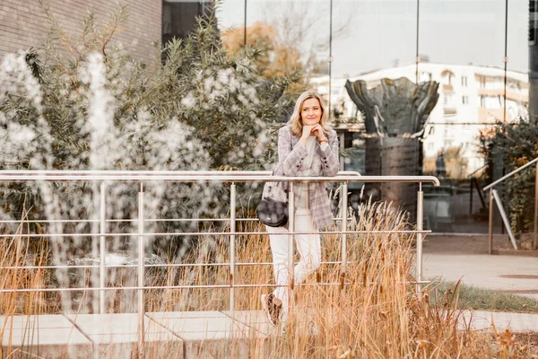 Atractiva mujer de mediana edad sonriente con chaqueta a cuadros y suéter gris con cuello enrollable de pie apoyando los codos en un puente de metal —  Fotos de Stock