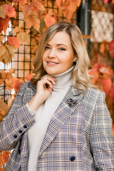 Donna sorridente di mezza età con i capelli lunghi in giacca a scacchi e maglione grigio roll-neck in posa toccante collo con mano. — Foto Stock