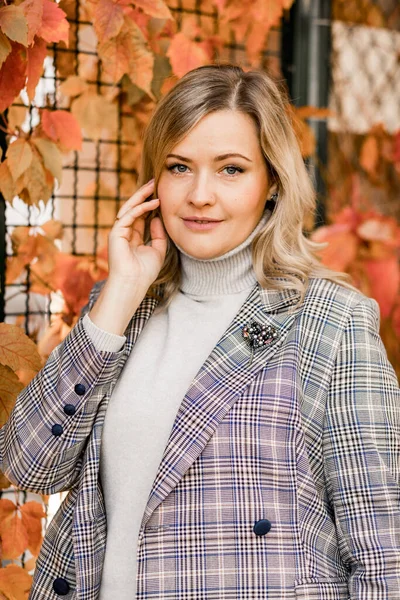 Bella donna sorridente di mezza età in giacca a scacchi e grigio roll-neck maglione posa titivating lunghi capelli biondi. — Foto Stock