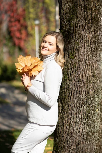 Donna sorridente di mezza età con lunghi capelli biondi ondulati in maglione roll-neck con mazzo di foglie cadute appoggiate sull'albero — Foto Stock