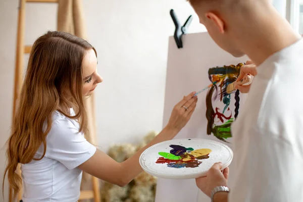 Young cute couple in white T-shirts stand painting absorbedly on canvas using watercolor palette, brushes near ladder. — Stock Photo, Image