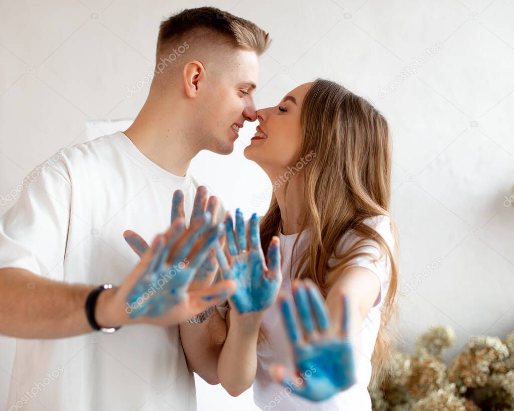 Smiling young pretty couple with fair hair stand closing eyes, nuzzling noses, showing hands covered with blue paint.