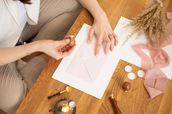 Foto recortada de manos de mujer haciendo artesanía sobre invitación de deshierbe, decorar adornar cinta rosa y cera de sello. Hobby. —  Fotos de Stock