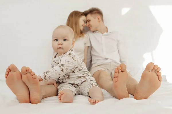 Retrato de jovem casal feliz olhando um para o outro em roupas brancas com bebê bebê querúbico criança sentada na cama. — Fotografia de Stock