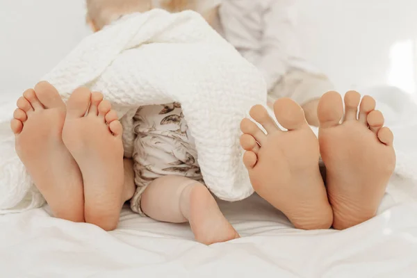 Acercamiento de los pies descalzos de la familia acostados en la cama con el bebé pequeño niño que está a cuatro patas cubierto con manta. —  Fotos de Stock