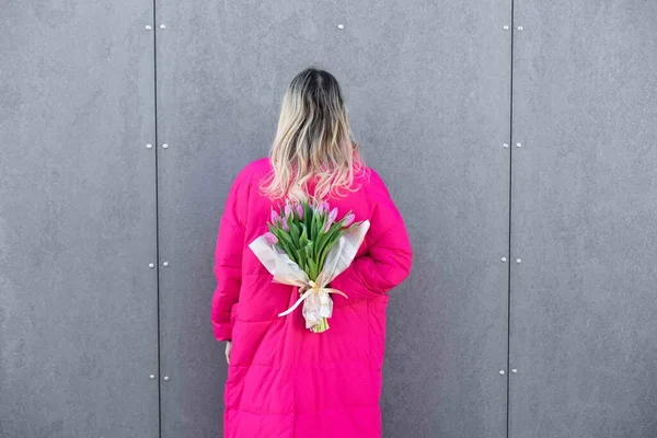 Porträtt av blond vacker kvinna i rosa jacka vistas nära metallvägg med rosa bukett tulpan blommor. Födelsedagsfest — Stockfoto
