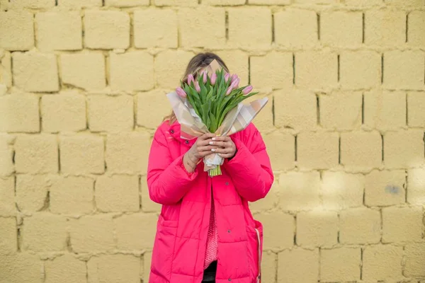Kvinna i rosa jacka vistas nära gul tegelvägg, håller rosa bukett tulpan blomma. Döljer ansiktet. Semesterfirande — Stockfoto