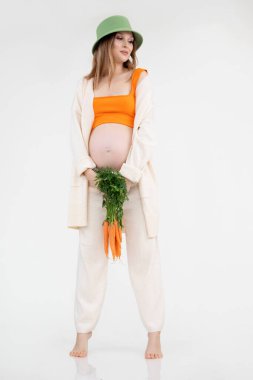 Çekingen, yalın ayak, gülümseyen, çekici bir kadının dikey fotoğrafı, yeşil şapka kalıyor, bir sürü havuç. Hamilelik tütsüsü.