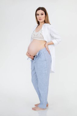 Çekici, kendine güvenen, çıplak ayaklı hamile bir kadının dikey fotoğrafı üstte, gömlek ve makyajlı bir pantolon. Karna dokunuyor..
