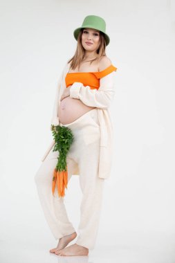 Parlak çıplak ayaklı, yeşil şapkalı, gülümseyen çekici bir kadının dikey fotoğrafı havuç demeti. Hamilelik epilasyonu.