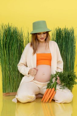 Çimlerin yanında yeşil şapkalı, içi çiğ havuçla dolu karnına bakan mutlu hamile kadının dikey hâli. Sarı görünüm