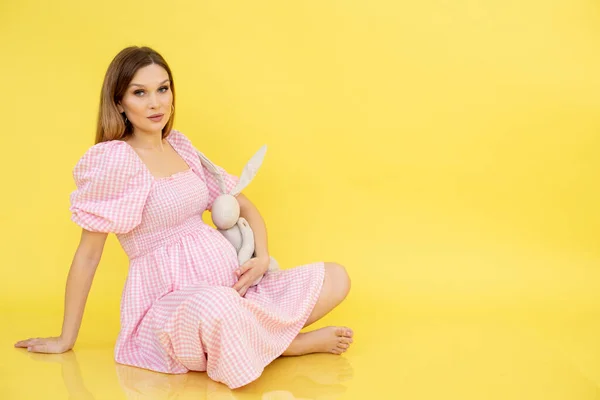 Blij zelfverzekerd kalmeren zwangere blootsvoets vrouw zittende vloer in roze jurk met konijn speelgoed. Wachten op baby meisje, ouderschap — Stockfoto