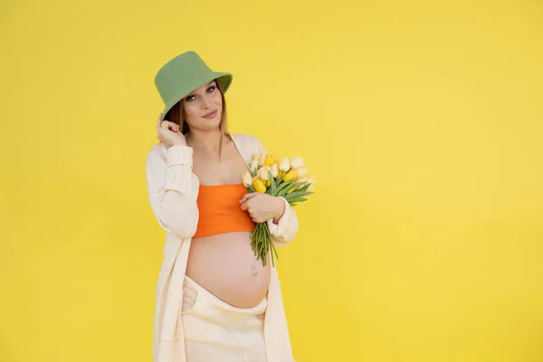 밝은 옷을 입고, 녹색 모자를 쓰고, 맨 배를 하고, 화려 한 튤립 꽃다발을 들고 있는 자신있는 임신부를 기쁘게 미소짓고 있다. 새 가게 — 스톡 사진