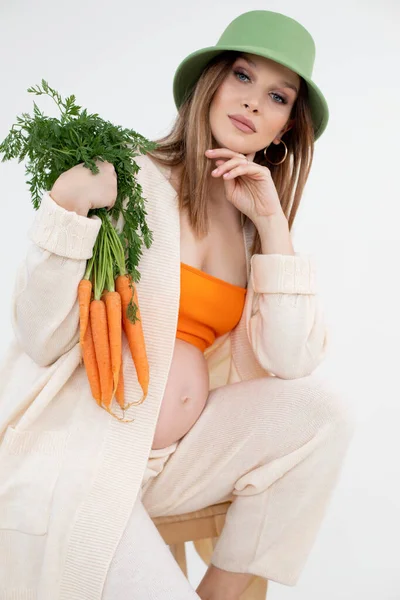 Vertical beauteous pregnant woman green hat, orange top and flax clothes, naked belly looking camera, orange carrots. — стоковое фото