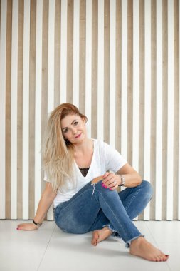 Gülümseyen orta yaşlı, güzel bir kadın. Uzun sarı saçlı, beyaz tişörtlü, mavi yırtık pantolonlu, bacak bacak üstüne atmış..