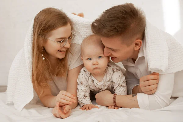 白いベッドスプレッドに横たわっている女性、男性と乳児の赤ちゃんの幸せな笑顔の家族。タオルを覆う建物. — ストック写真