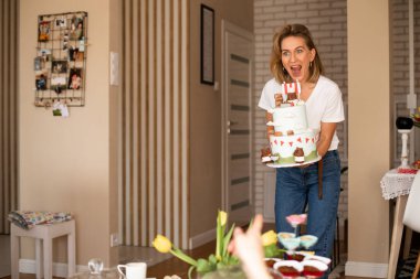 Ayakta duran ve büyük bir doğum günü pastası tutan kadın. Annem masaya yemek getiriyor, çocukların doğum günü için beyaz pasta..