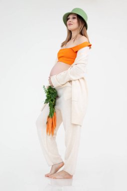 Uzun siyah saçlı çekici hamile kadın yeşil kova şapka takıyor, ayakta duruyor, karnını tutuyor ve havuç yiyor..