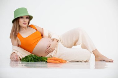 Uzun siyah saçlı, yeşil kova şapkalı, havuç demetinin yanında yatan güzel hamile kadının portresi..