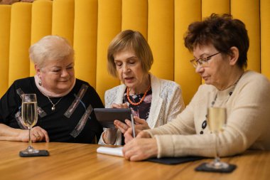 Üç tatlı, özümsenmiş yaşlı kadın hesap makinesine kalemle işaret ederek masada oturuyor ve deftere notlar alıyor..