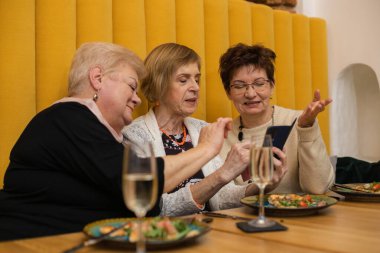 Üç tane gülümseyen yaşlı kadın cep telefonlarına bakar, tahta masada pizza ve şampanya kadehleriyle otururlar..